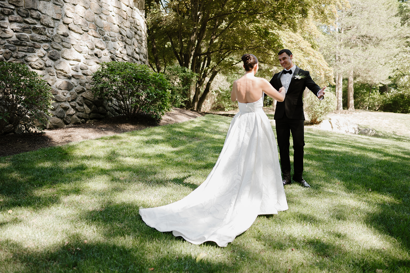 Groom reacting with a joyful smile during a first look on the lawn by a stone wall.